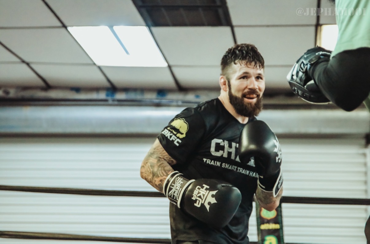 Dave Mundell smiling during pad work in a black Chute Boxe shirt and BKFC cap